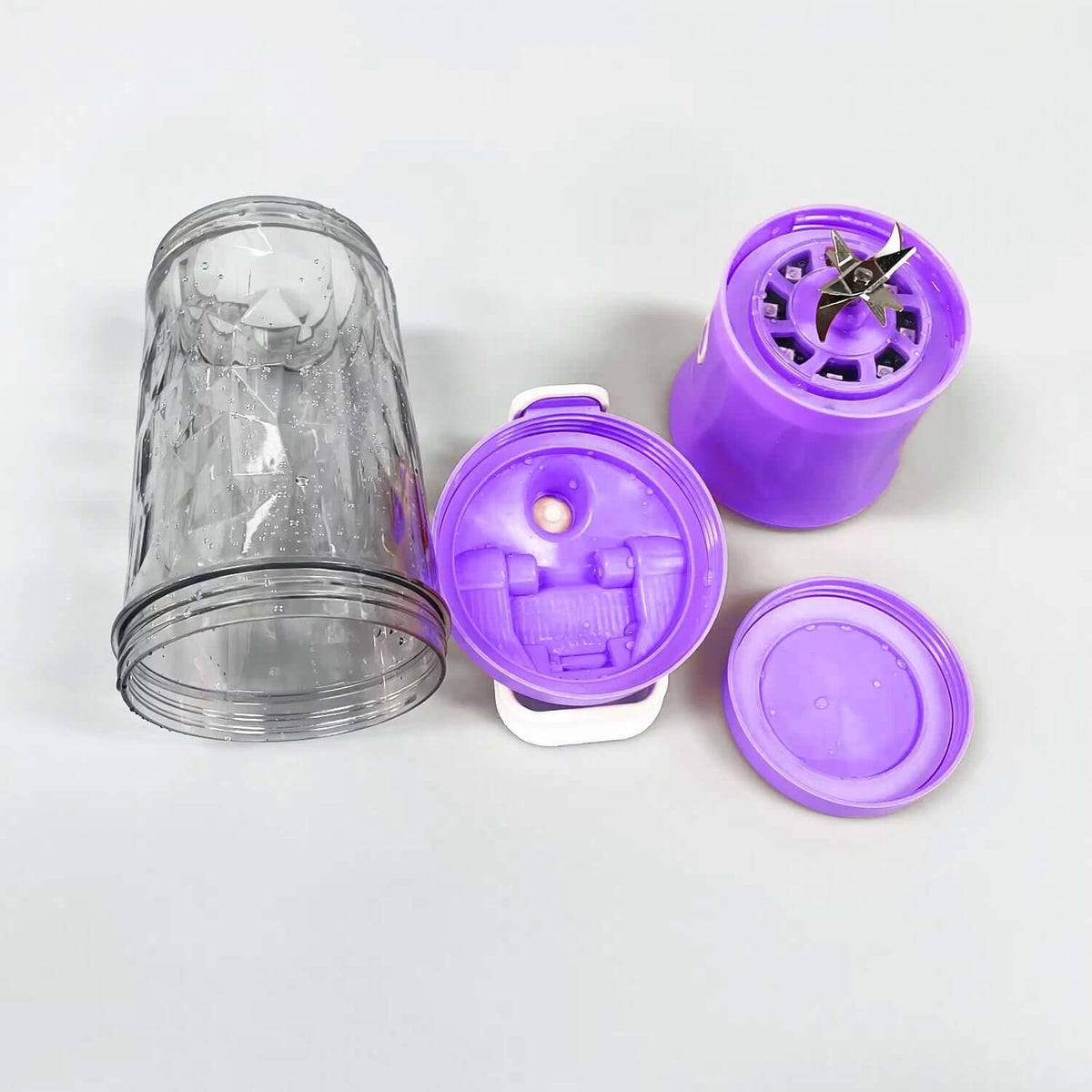 Portable electric juicer blender parts including purple cup and stainless steel blades on a gray surface.