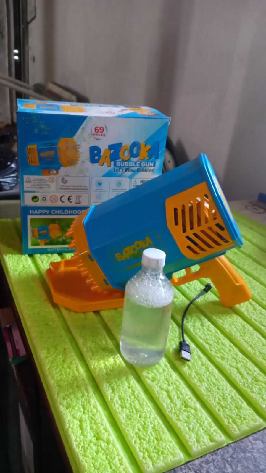 69-Hole Rechargeable Bubble Gun for Kids (Rocket Boom) with bottle and charging cable on a table.